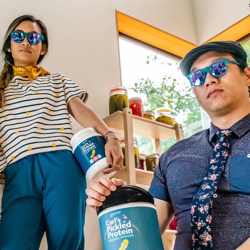 A man and woman wearing teal circle-shaped sunglasses with round reflective purple lenses stand beside pickled vegetables.