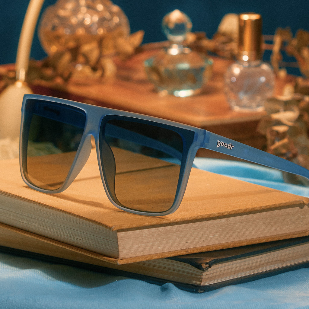 A pair of oversized, dusty blue Fly G sunnies sits atop vintage books, with dried blooms and perfume in the background.