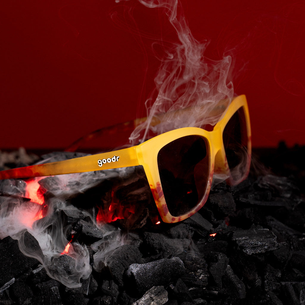 Three-quarter angle view of translucent yellow-orange goodr sunglasses on top of smoking coals.