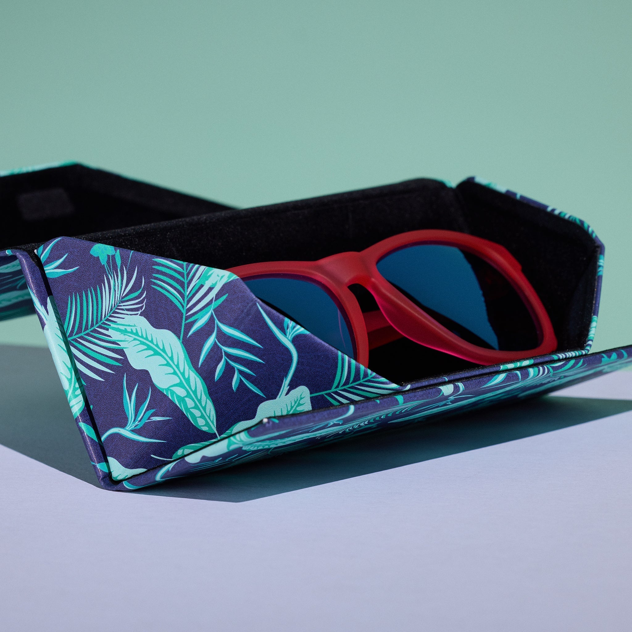 Close-up view of the Enter the Fold navy and teal tropical-print sunglasses case, shown open, with a pair of red sunnies inside.