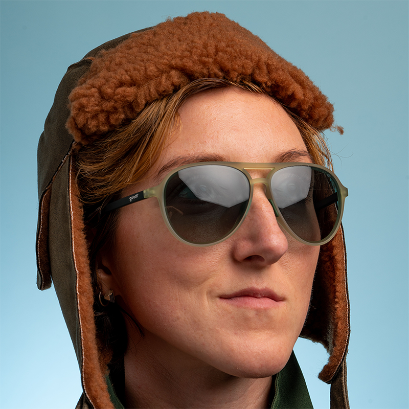 A woman wearing a brown fuzzy aviator cap looks out confidently, wearing cadet green aviators with green gradient lenses.