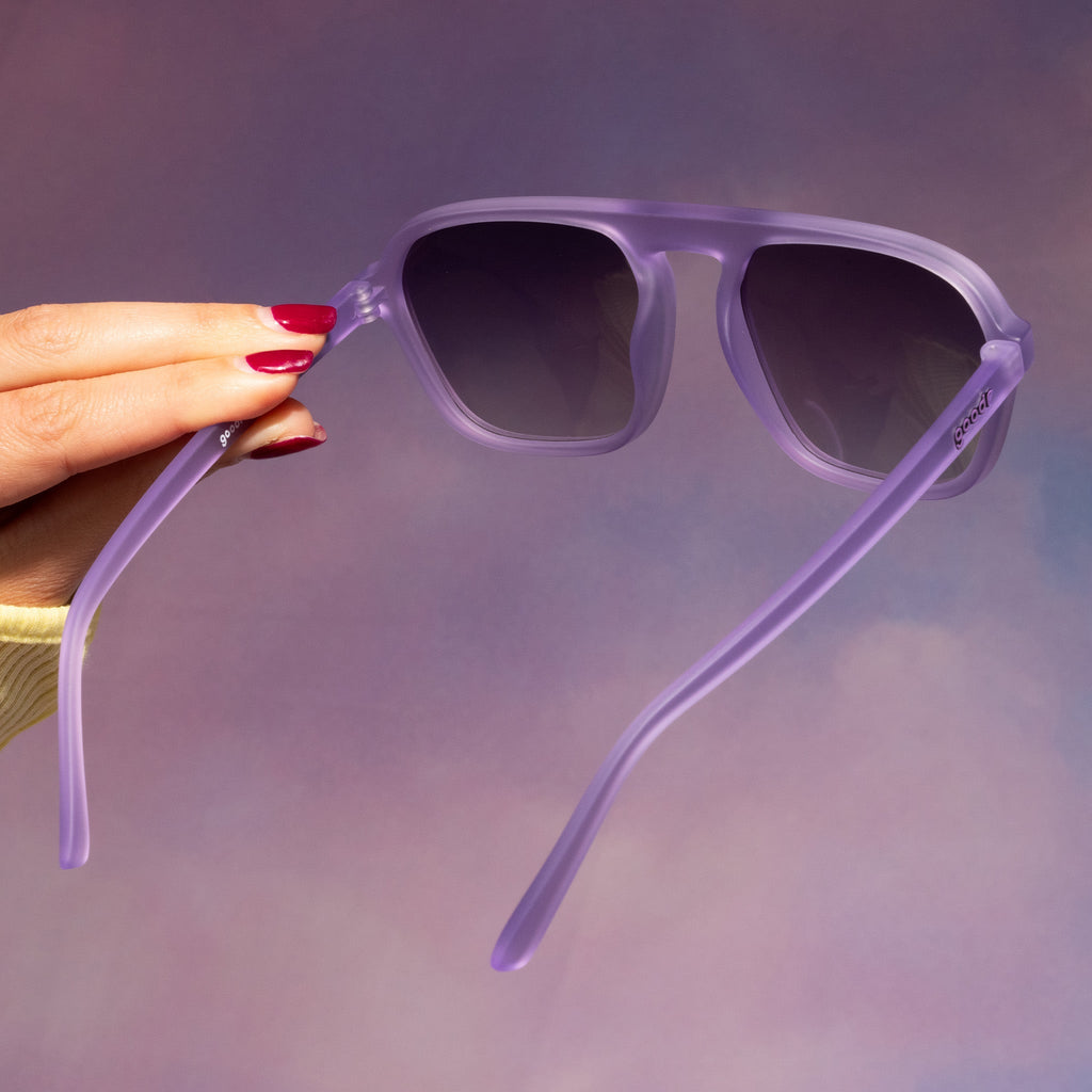 A manicured hand with red nails holds up a pair of translucent pale purple Retro G sunglasses against a pale purple backdrop.