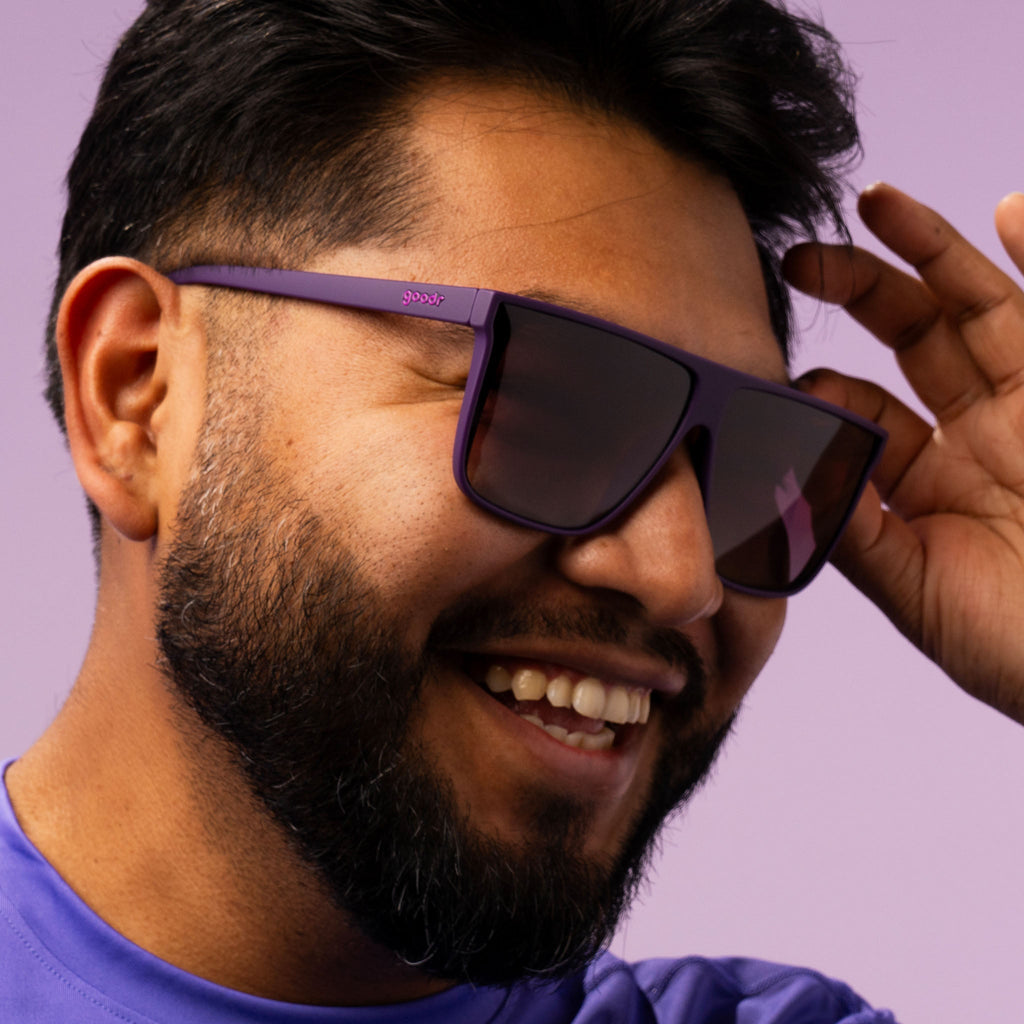 A closeup of a smiling man wearing a purple shirt playfully tipping his oversized dark purple sunglasses.