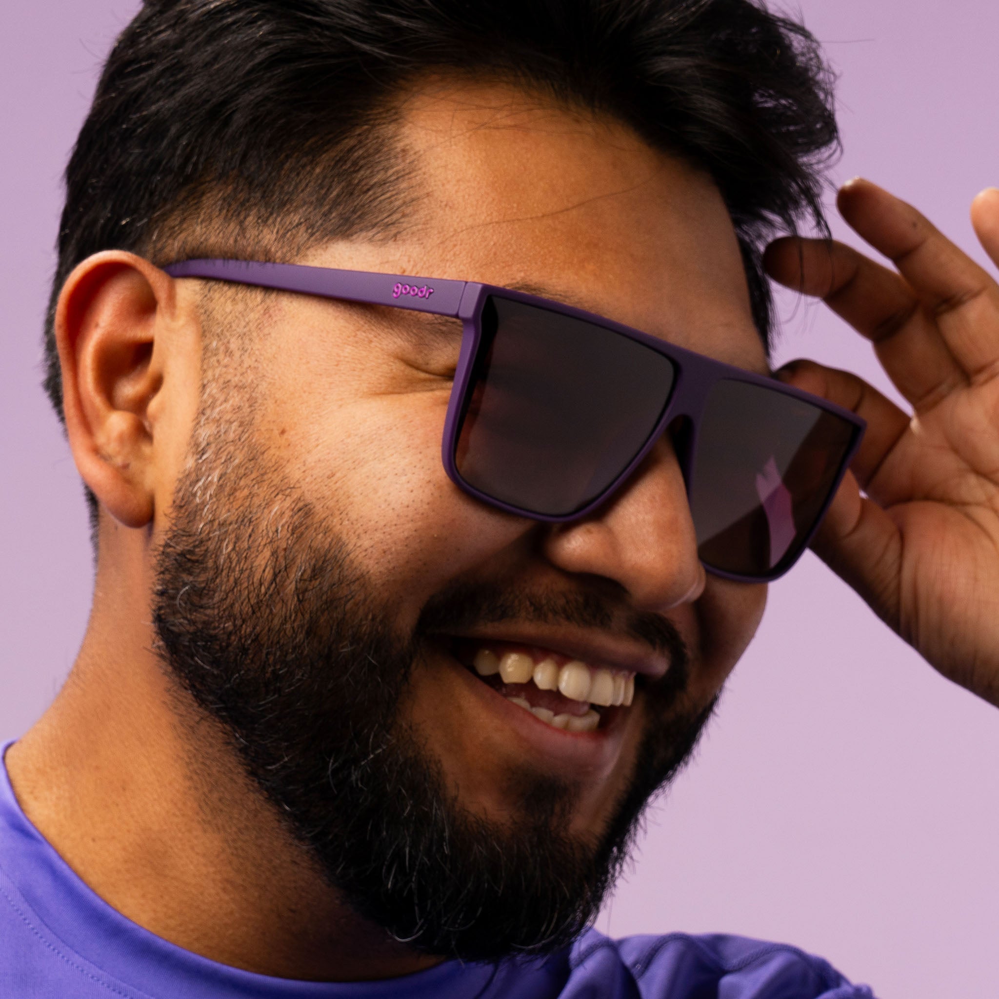 A closeup of a smiling man wearing a purple shirt playfully tipping his oversized dark purple sunglasses.