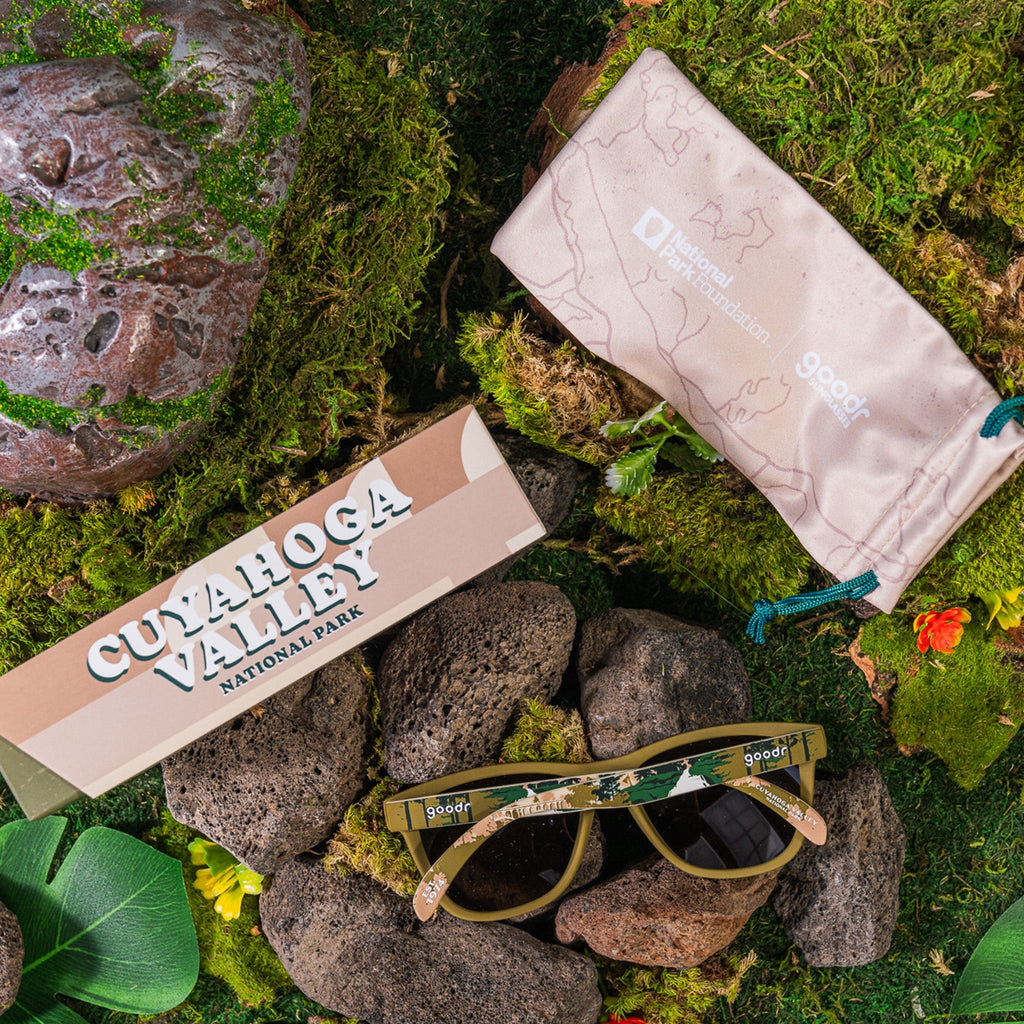 A pair of olive green "Cuyahoga Valley" goodr sunglasses, featuring nature-inspired designs across the temples, sitting on some rocks next to their box and a beige case.