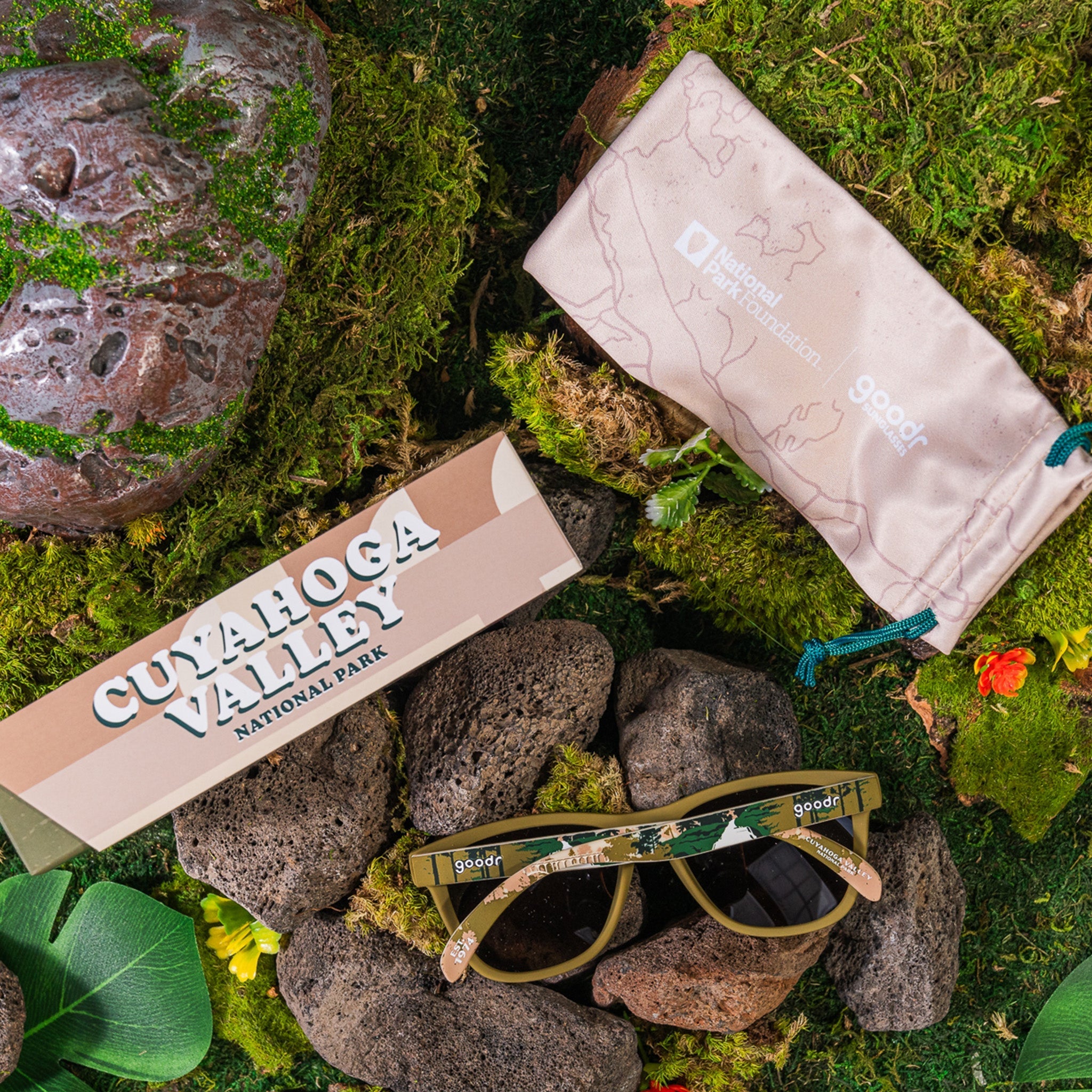 A pair of olive green "Cuyahoga Valley" goodr sunglasses, featuring nature-inspired designs across the temples, sitting on some rocks next to their box and a beige case.