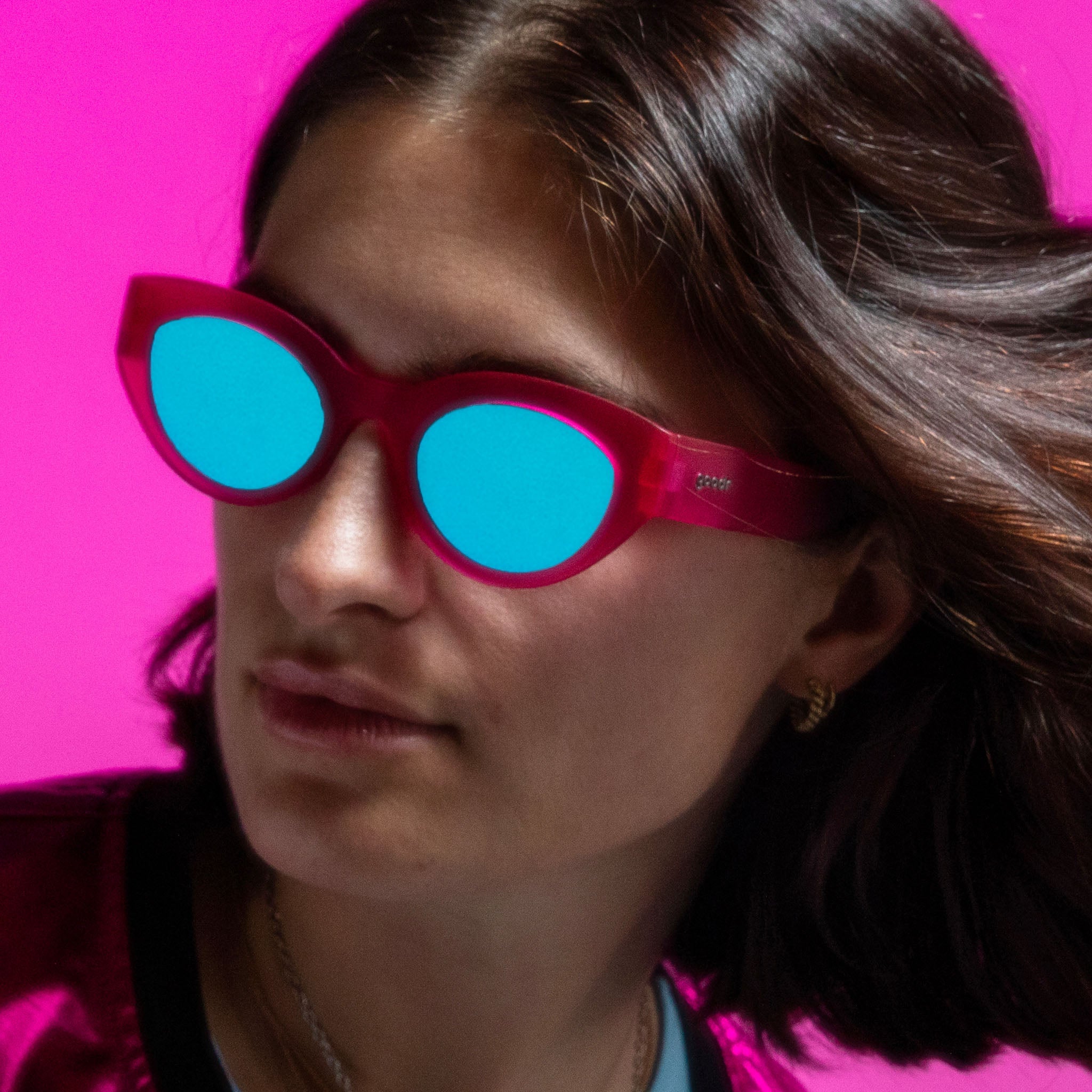 A model with coppery feathered hair wears a pair of translucent hot pink cat-eye frames with striking blue reflective lenses.