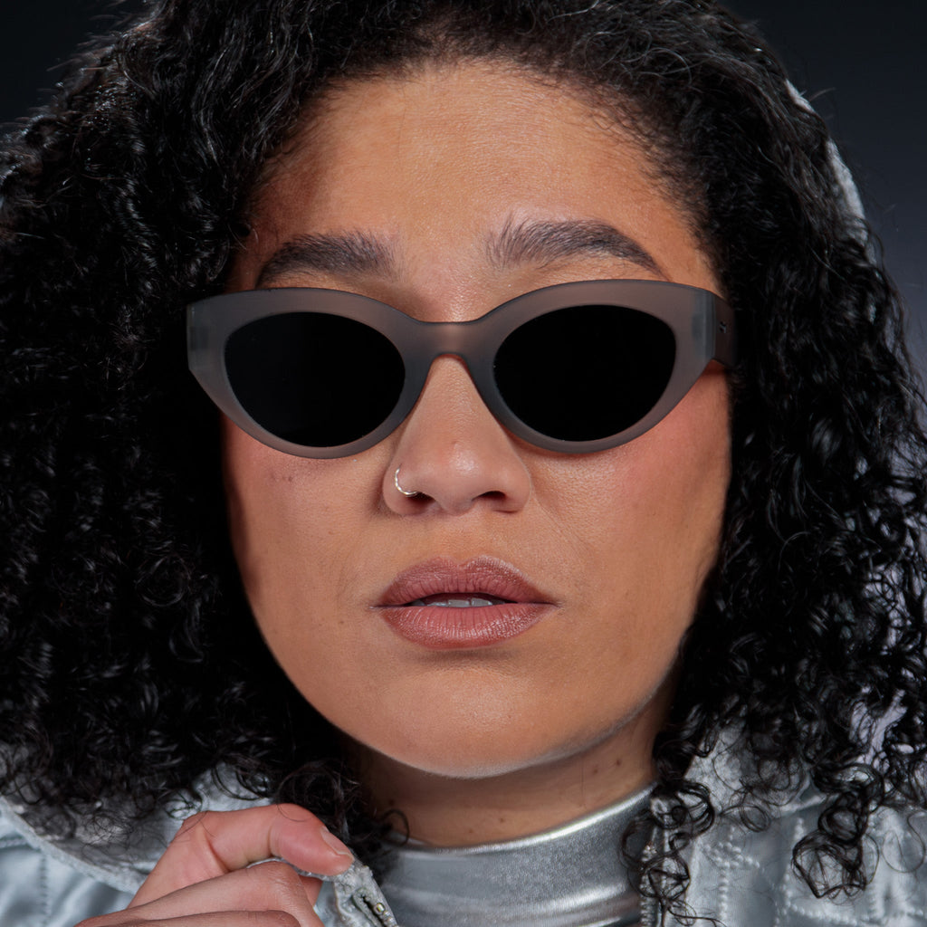 Close-up of a woman posing while wearing translucent dark gray, retro-inspired angled cat-eye sunglasses with black non-reflective lenses.