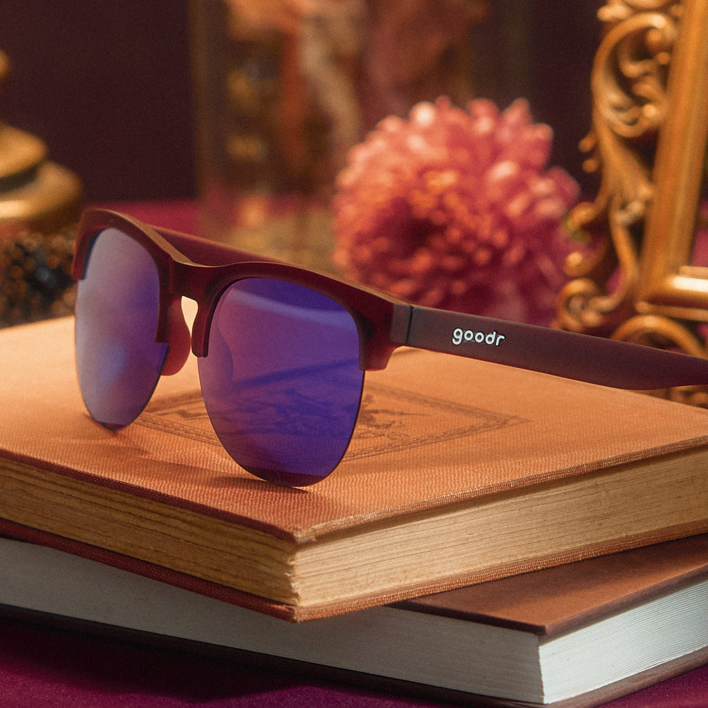 Three-quarter angle view of translucent purple half-rim sunglasses with purple reflective lenses on top of stacked books.