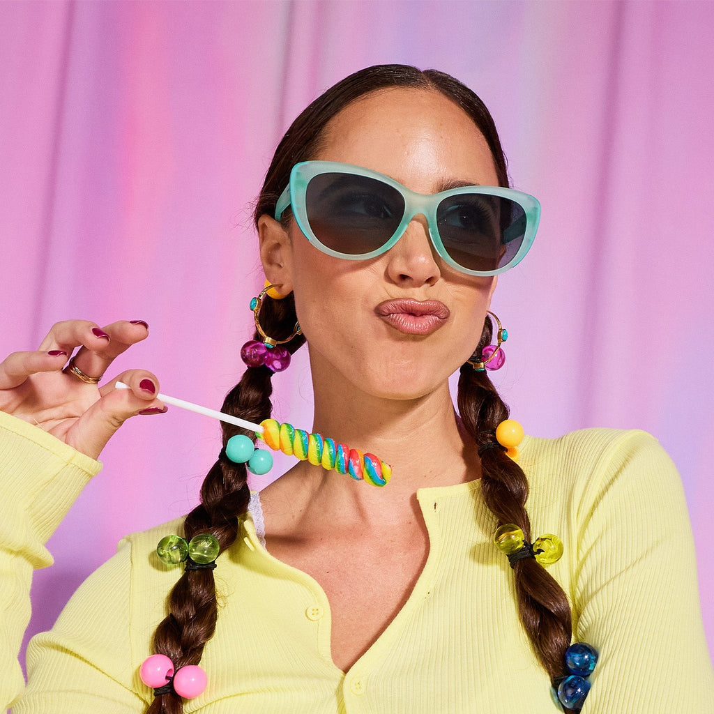 A woman with dark pigtails models pale-green cat-eye sunglasses against a candy pink backdrop, holding a rainbow lollipop.