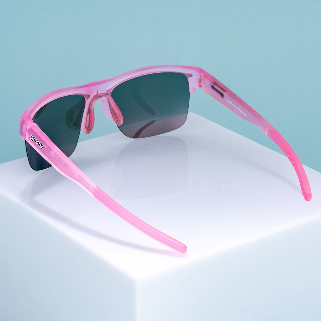 Three-quarter back angle view of half-rim translucent pink sunglasses with pink reflective polarized lenses. The arms are extended, showcasing the goodr logo on the outer left arm, an adjustable light pink silicone nose piece, and pink silicone grips on the inner arms.