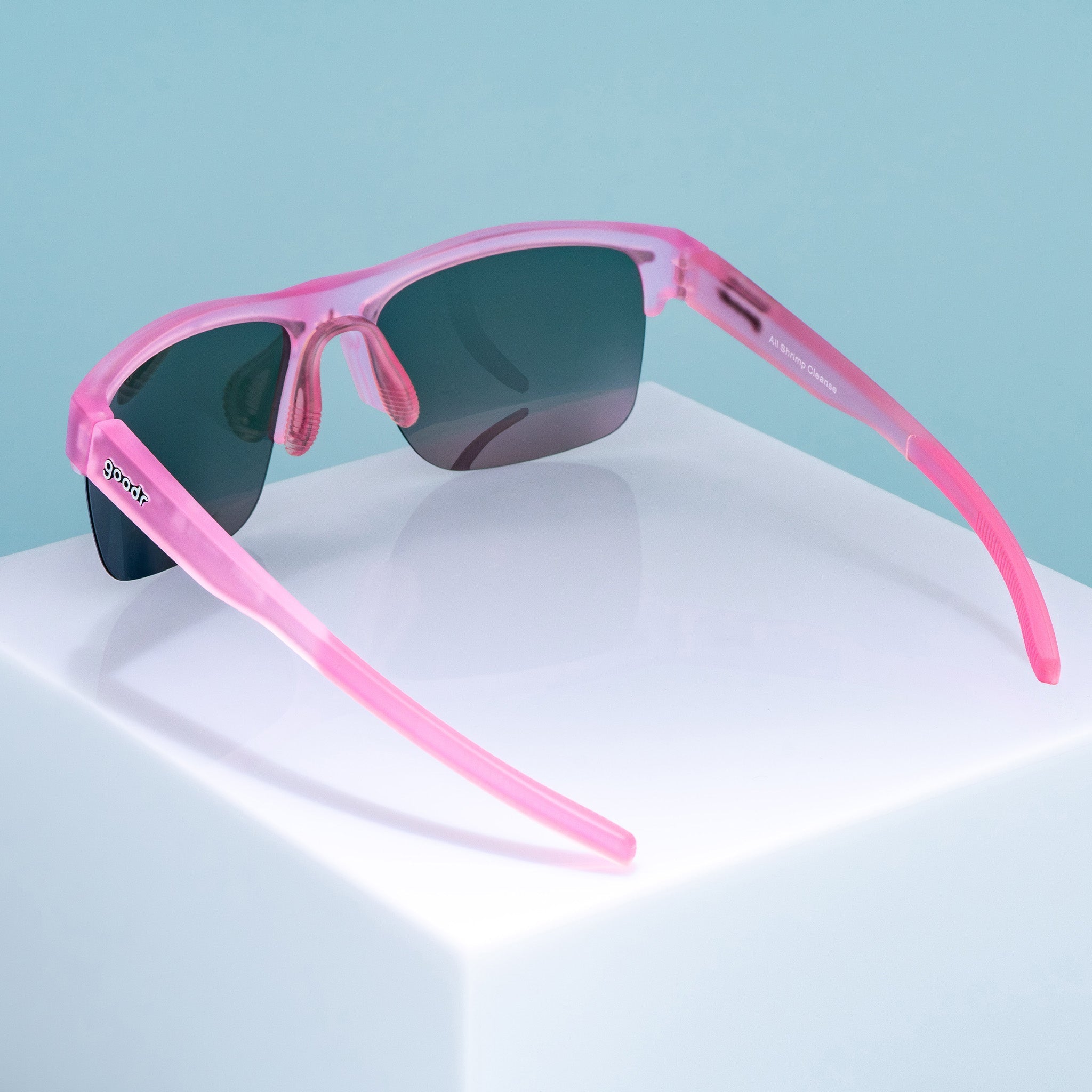 Three-quarter back angle view of half-rim translucent pink sunglasses with pink reflective polarized lenses. The arms are extended, showcasing the goodr logo on the outer left arm, an adjustable light pink silicone nose piece, and pink silicone grips on the inner arms.