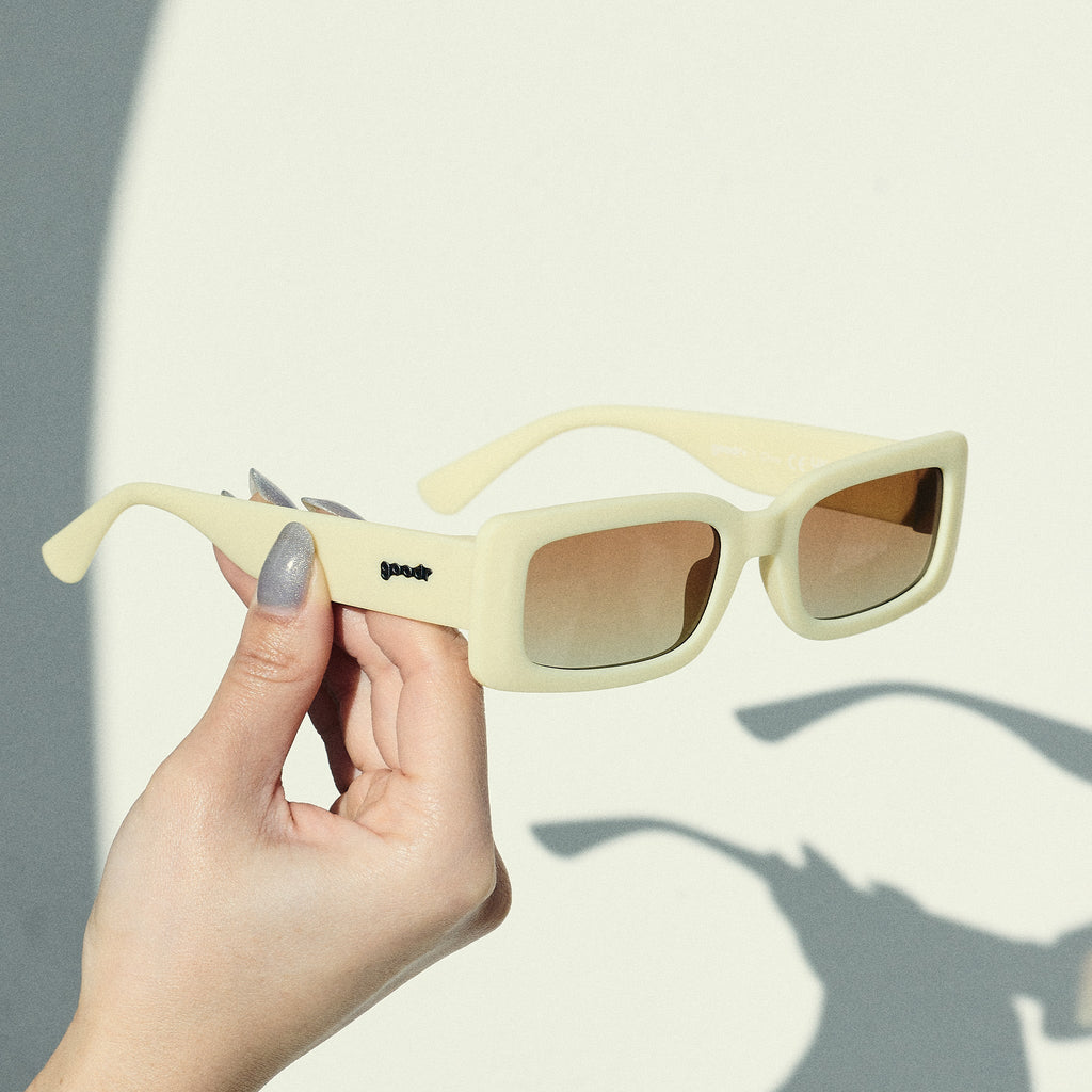 A manicured hand holds up a pair of yellow rectangular sunglasses with brown-tinted polarized lenses in front of an off-white backdrop.