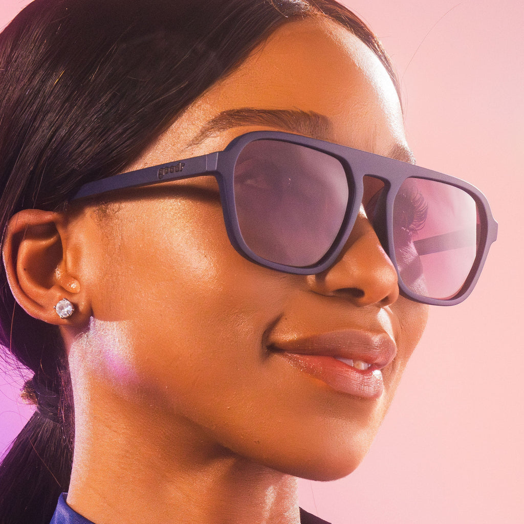 A close-up of a woman wearing retro aviators sunglasses with dark frames and lenses. She has sleek, pulled-back hair and is accessorized with small, sparkling stud earrings.