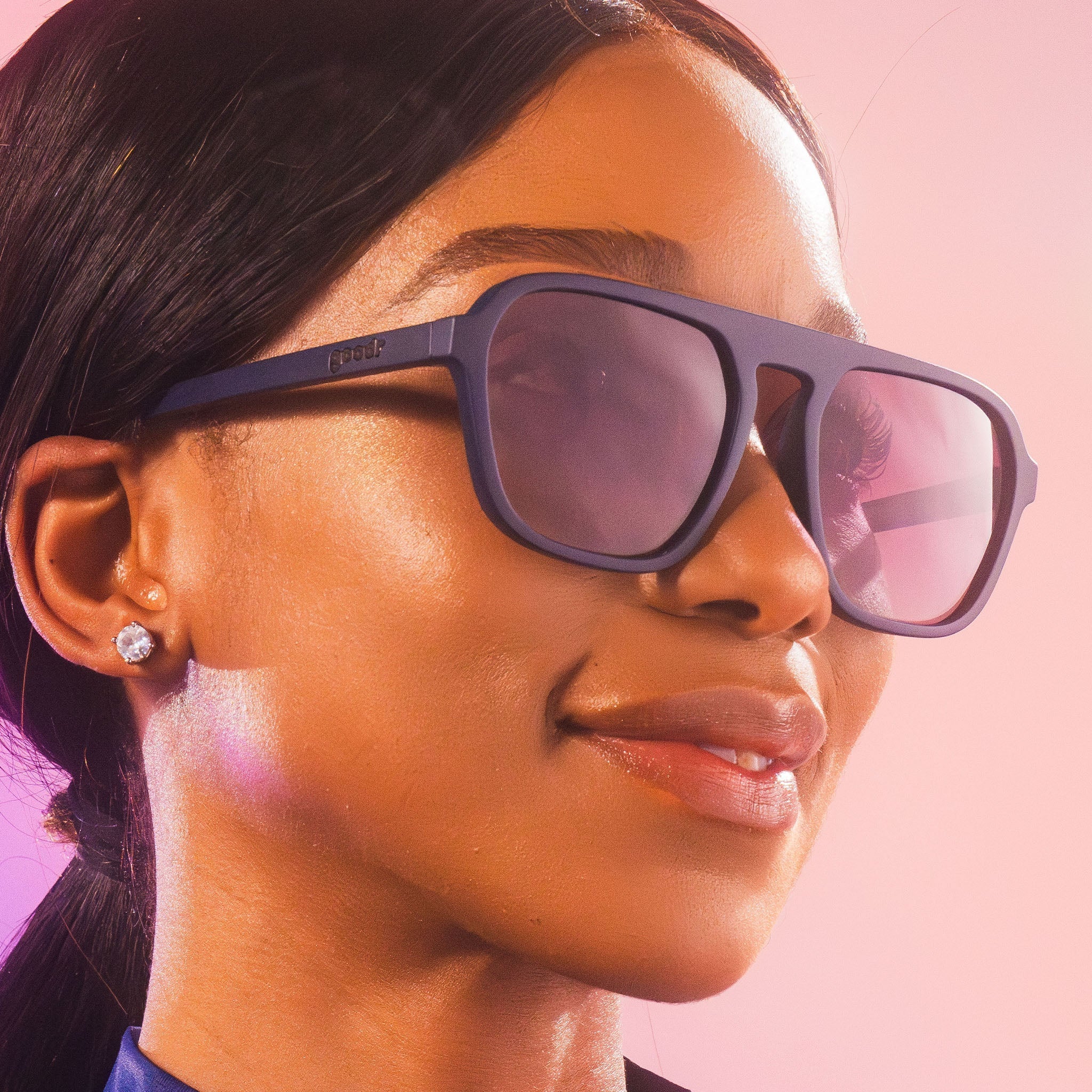 A close-up of a woman wearing retro aviators sunglasses with dark frames and lenses. She has sleek, pulled-back hair and is accessorized with small, sparkling stud earrings.