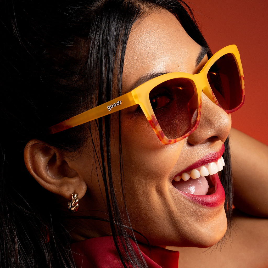 A woman looks over her shoulder while smiling and wearing translucent yellow-orange goodr sunglasses.